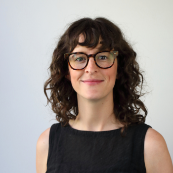 Portrait of a woman with curly hair and glasses, smiling, wearing a sleeveless black top.
